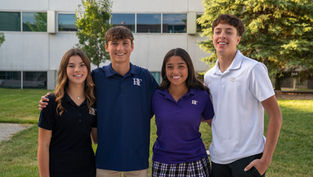 Four smiling students in colored polo shirts and plaid skirt, standing outdoors in front of a school building with trees in the background.