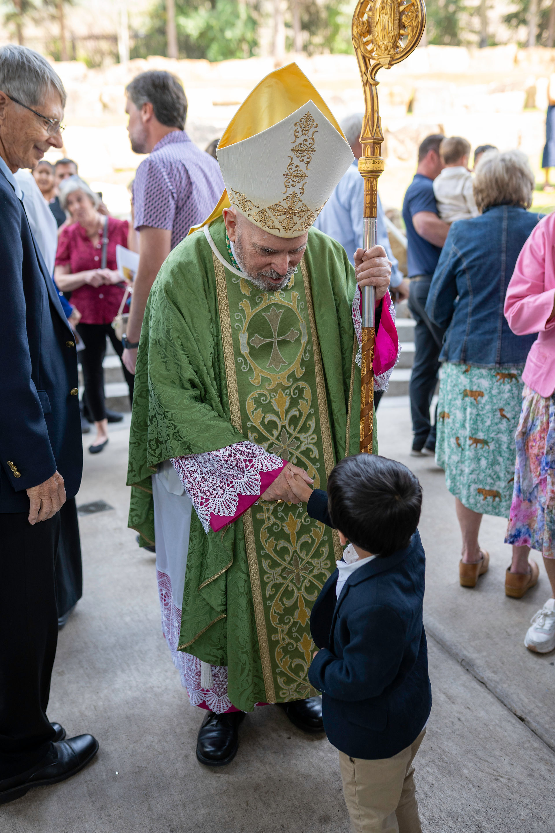 A bishop in green and gold vestments, wearing a mitre and holding a crosier, shakes hands with a young boy in formal attire. Several people are visible in the background in an outdoor setting.