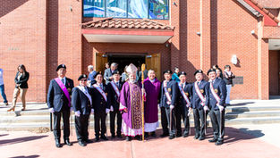 A group of men in ceremonial attire with a bishop in purple vestments pose outside a red brick church, under a vibrant stained glass window.