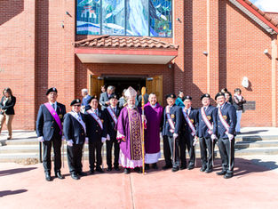 A group of men in ceremonial attire with a bishop in purple vestments pose outside a red brick church, under a vibrant stained glass window.