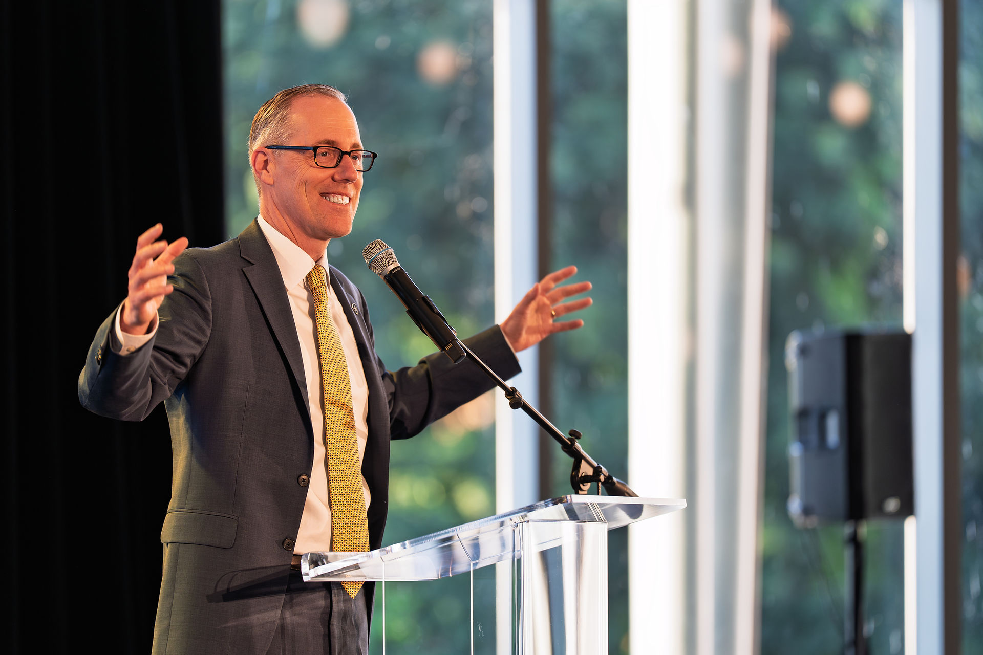 Smiling man in a suit and yellow tie speaks at a clear podium. He gestures openly, with greenery visible through large windows behind him.