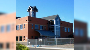 Brick building with large windows and a small tower stands against a clear blue sky, surrounded by a metal fence and playground.