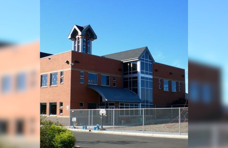 Brick building with large windows and a small tower stands against a clear blue sky, surrounded by a metal fence and playground.
