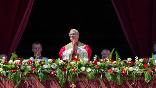 A religious figure in red and white robes stands on a flower-adorned balcony with hands in prayer, surrounded by men and red curtains.