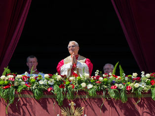 A religious figure in red and white robes stands on a flower-adorned balcony with hands in prayer, surrounded by men and red curtains.