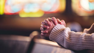 Hands clasped in prayer, resting on a wooden pew. A cozy sweater and blurred colorful stained glass background convey a peaceful mood.
