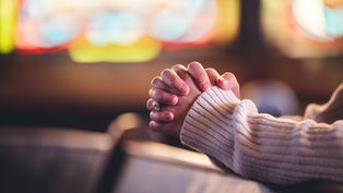 Hands clasped in prayer, resting on a wooden pew. A cozy sweater and blurred colorful stained glass background convey a peaceful mood.