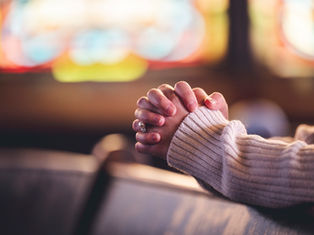 Hands clasped in prayer, resting on a wooden pew. A cozy sweater and blurred colorful stained glass background convey a peaceful mood.