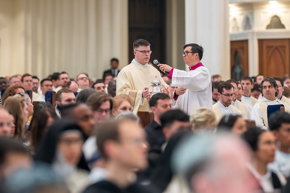 Priests in ceremonial robes participate in a religious service. One holds a vessel, while another speaks into a microphone. Formal setting.
