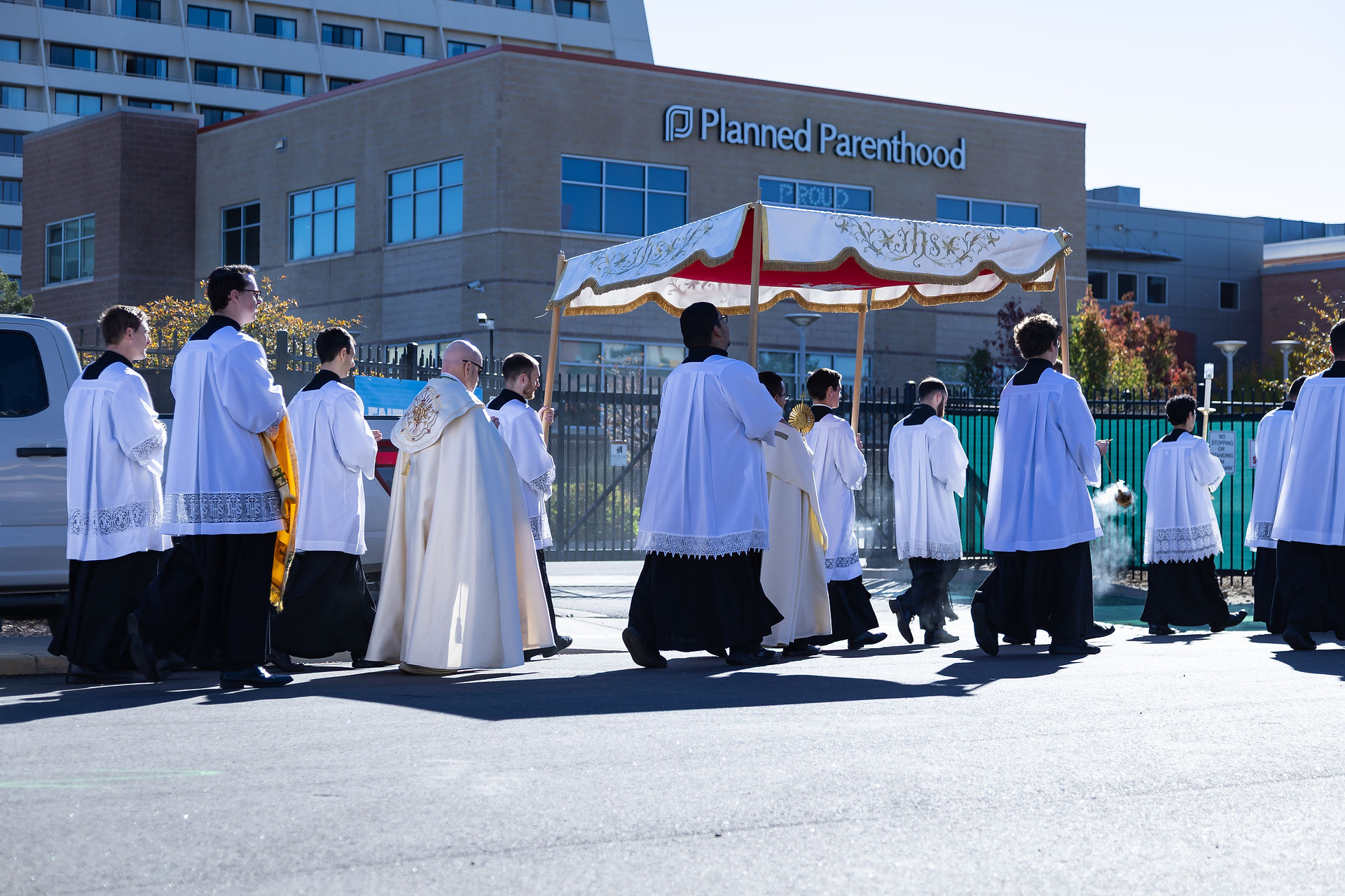 Priests in white robes participate in a religious procession outside a Planned Parenthood building. A golden canopy is carried above.