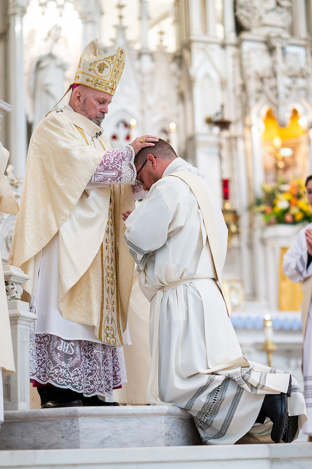 A bishop prays over a kneeling man in a white robe inside an ornate church. The scene is solemn with intricate gold details and floral decor.