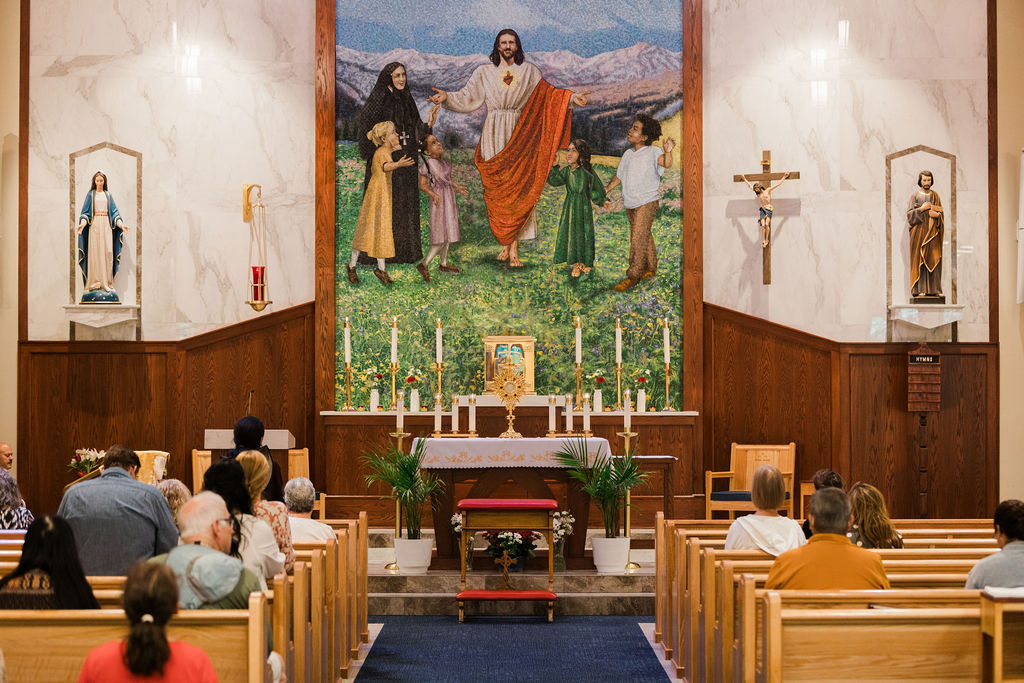 Church sanctuary with worshippers facing altar, crucifix and statues, and a mural of Jesus with children above.