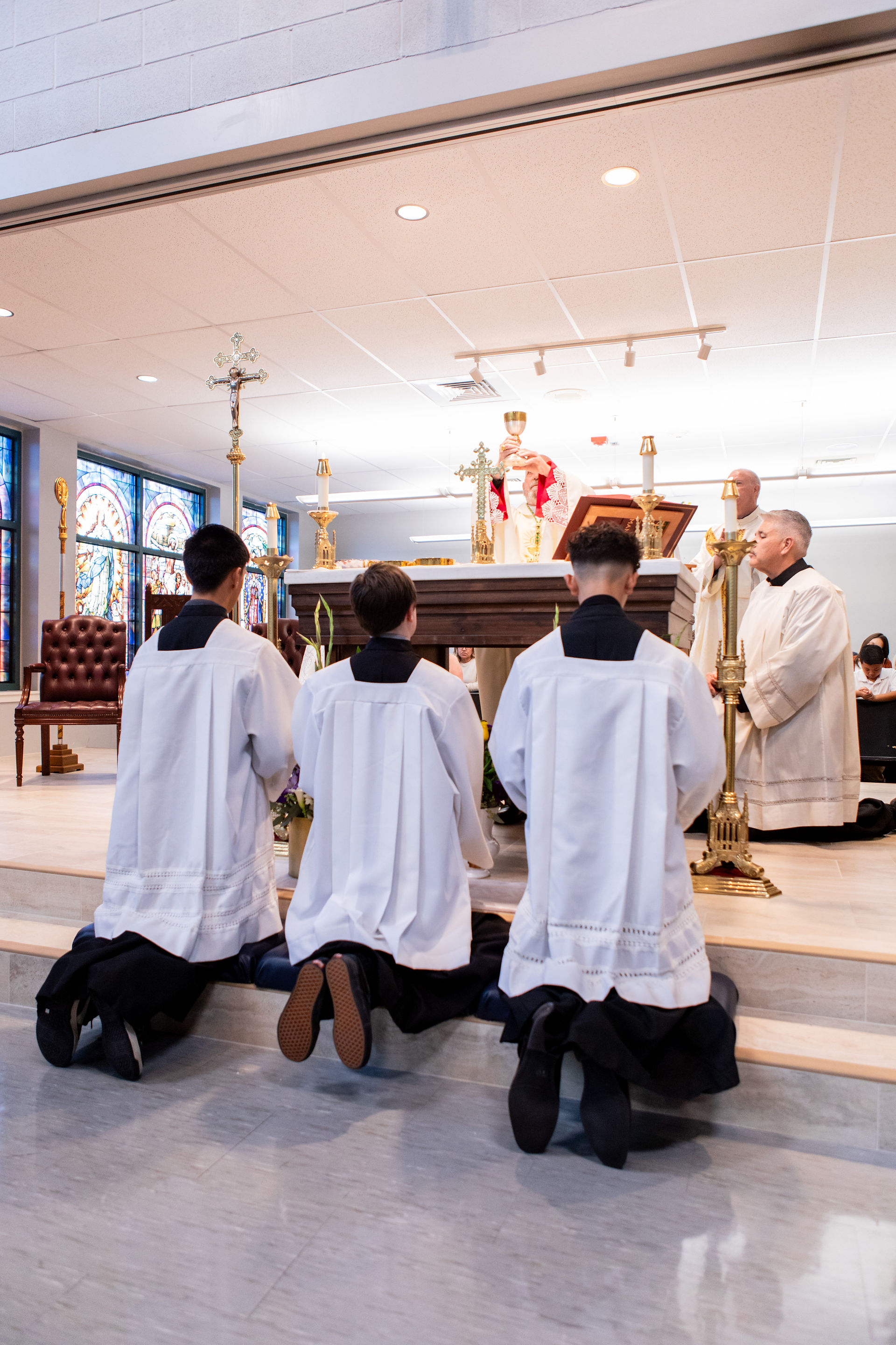 Four people in white robes kneel before an altar during a church service. Stained glass windows and a solemn atmosphere are present.