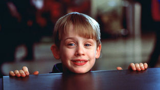 Young boy smiling over a table in a busy background, wearing a dark outfit. His hands grip the table's edge, creating a cheerful mood.