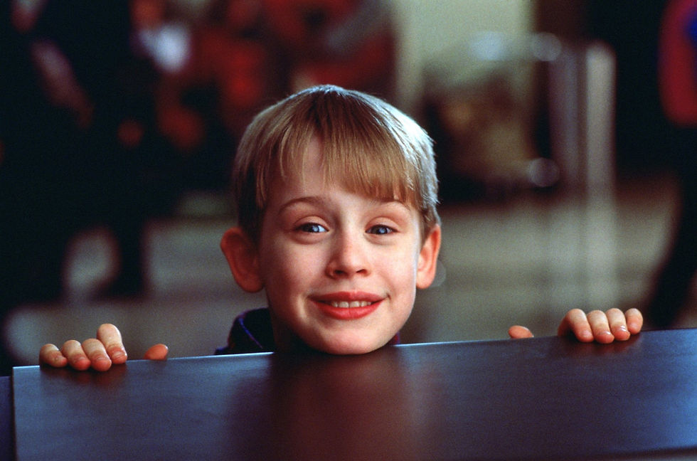 Young boy smiling over a table in a busy background, wearing a dark outfit. His hands grip the table's edge, creating a cheerful mood.