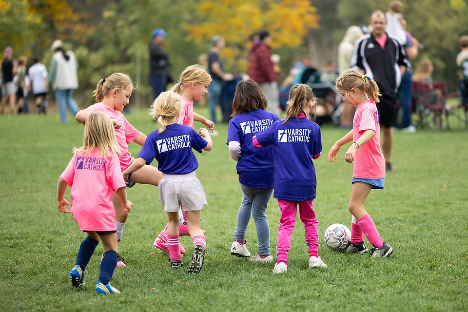 Young girls in pink and blue Varsity Catholic jerseys play soccer on a grassy field, with a ball nearby and spectators behind.