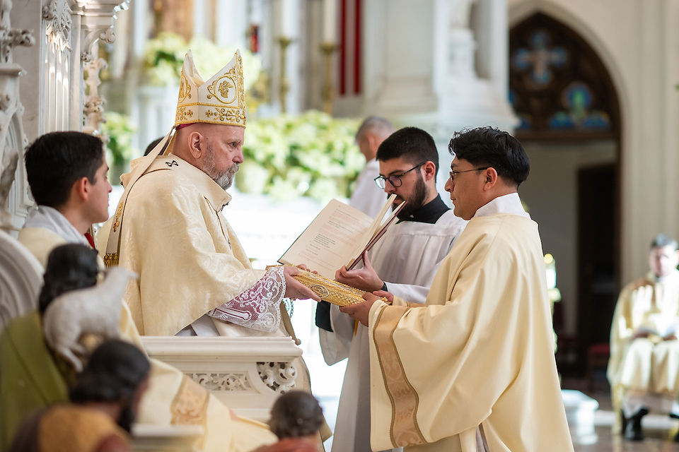 A bishop in ornate attire conducts a ceremony with clergy holding books, set in a richly decorated church with sunlight filtering in.
