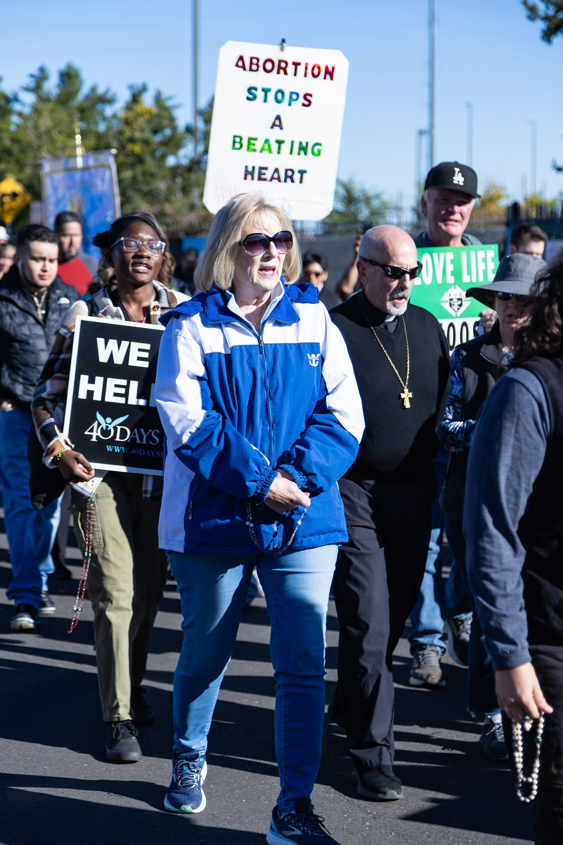 People at a demonstration hold signs: "Abortion stops a beating heart," "We help," and "Love life." They walk outdoors in a sunny setting.