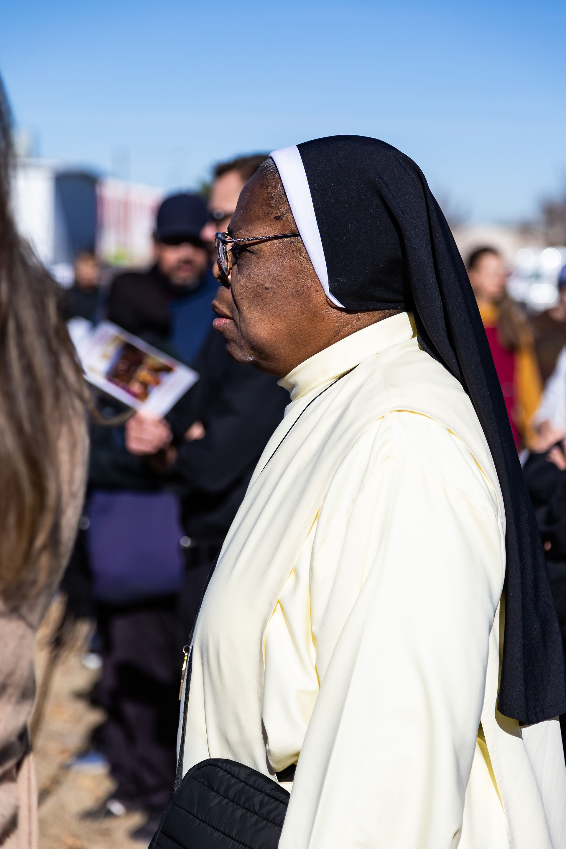 Nun in a white habit and black veil stands outdoors, surrounded by people. Clear blue sky in the background conveys a peaceful mood.