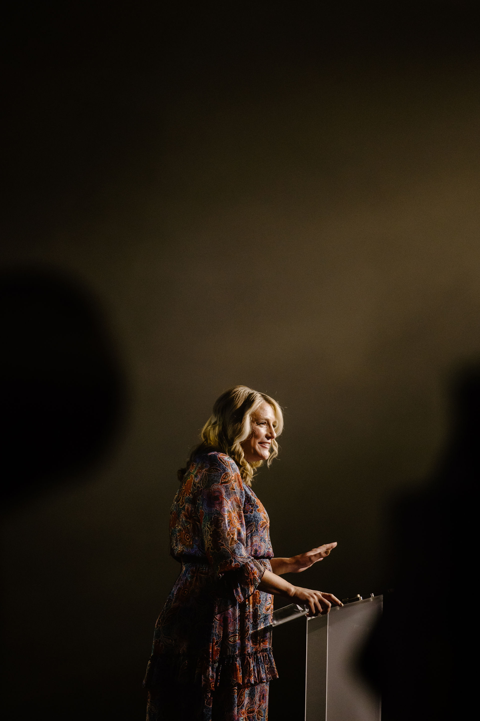 Woman with blonde hair in a patterned dress stands behind a podium, smiling against a dark, neutral background. She seems content.