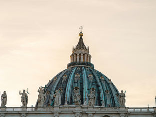 St. Peter's Basilica dome at sunset against a pale sky, flanked by statues. A serene and majestic architectural scene.