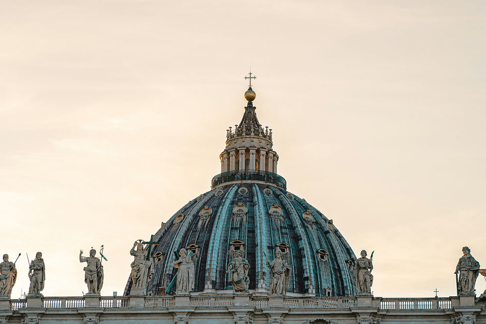 St. Peter's Basilica dome at sunset against a pale sky, flanked by statues. A serene and majestic architectural scene.