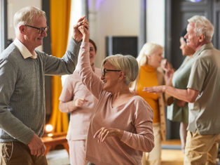 Elderly couple dancing joyfully indoors, wearing casual clothes. Other people chat in the background. Warm lighting and a cheerful atmosphere.