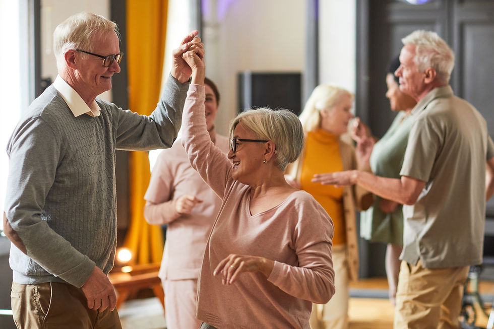 Elderly couple dancing joyfully indoors, wearing casual clothes. Other people chat in the background. Warm lighting and a cheerful atmosphere.
