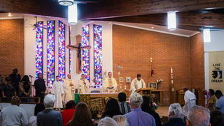 Priests conduct a service in a church with stained glass windows. Congregation watches. Banner reads "Live the Dream." Brick walls.