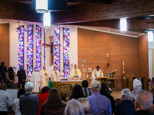 Priests conduct a service in a church with stained glass windows. Congregation watches. Banner reads "Live the Dream." Brick walls.
