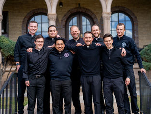 Nine men in clerical attire smile, posing together outdoors in front of an old building with arches. The mood is cheerful and relaxed.