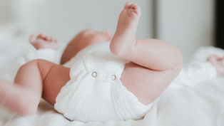 Baby lying on a white bed, wearing a white onesie, with legs playfully raised. Soft, calm atmosphere. No visible text.