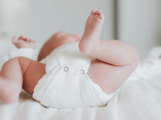 Baby lying on a white bed, wearing a white onesie, with legs playfully raised. Soft, calm atmosphere. No visible text.