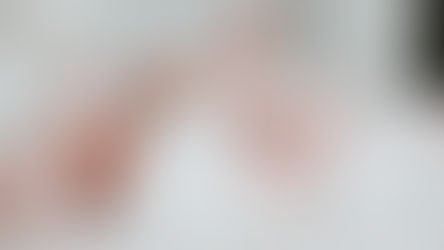 Baby lying on a white bed, wearing a white onesie, with legs playfully raised. Soft, calm atmosphere. No visible text.