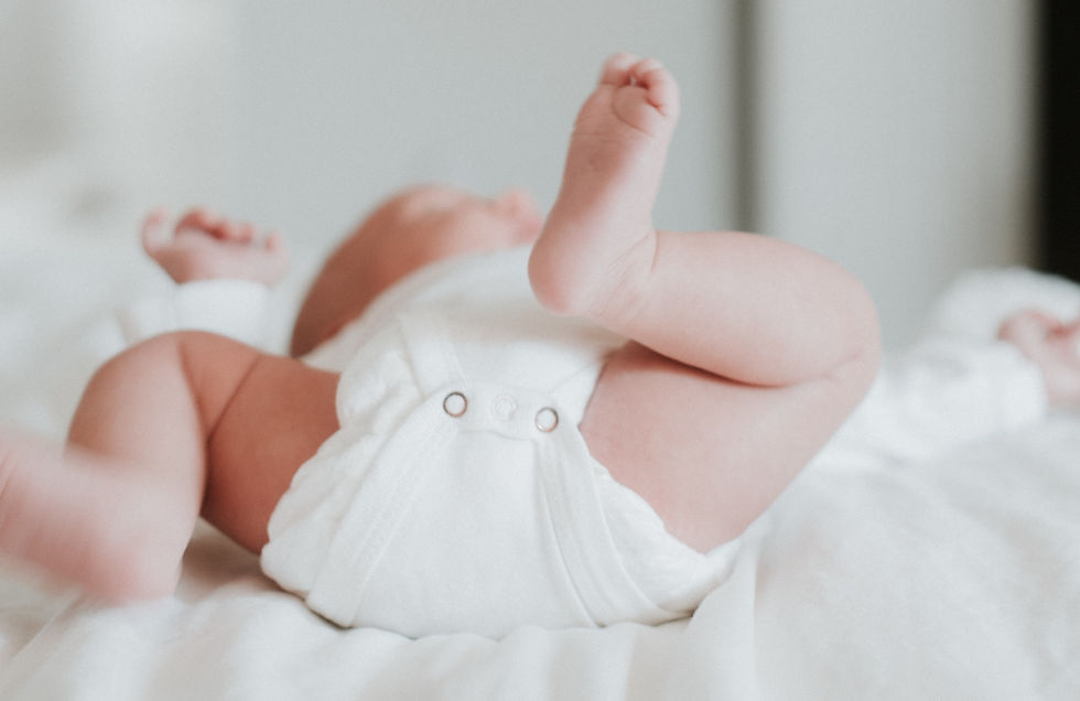 Baby lying on a white bed, wearing a white onesie, with legs playfully raised. Soft, calm atmosphere. No visible text.