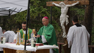 Outdoor Catholic Mass with bishop in green vestments holding a chalice beside a crucifix, under a white canopy in a forest.
