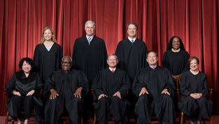 Seven Supreme Court justices in black robes pose before red curtains, seated and standing in a formal group portrait.