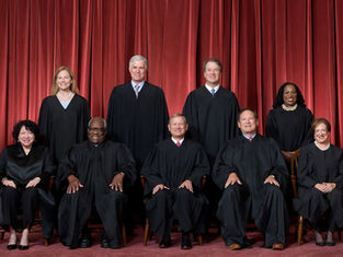 Seven Supreme Court justices in black robes pose before red curtains, seated and standing in a formal group portrait.