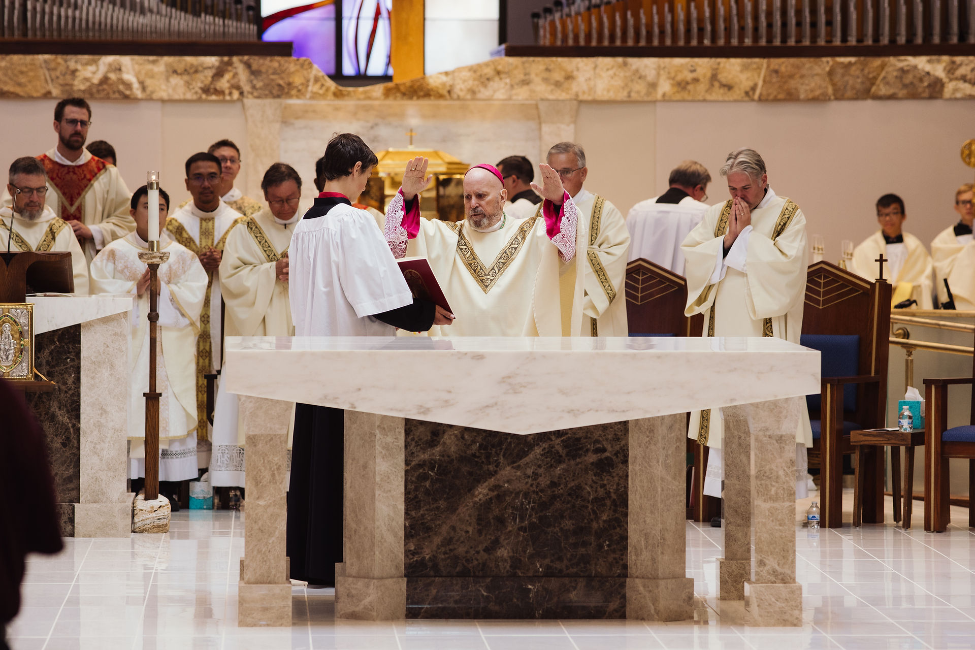 Clergy in ornate robes stand around an altar during a religious ceremony. One man raises hands in blessing. Stained glass in background.