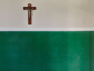 Crucifix on wall above green chalkboard with faint writing. Classroom setting with muted light and a contemplative, peaceful mood.
