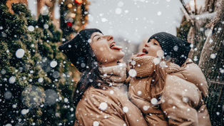 Two people in winter coats and hats catching snowflakes on their tongues. Snow falls against a background of evergreen trees. Joyful mood.