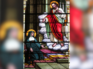 Stained glass depicting a haloed figure in red and green robes, standing on clouds, gesturing to a kneeling, haloed woman in a dark habit.