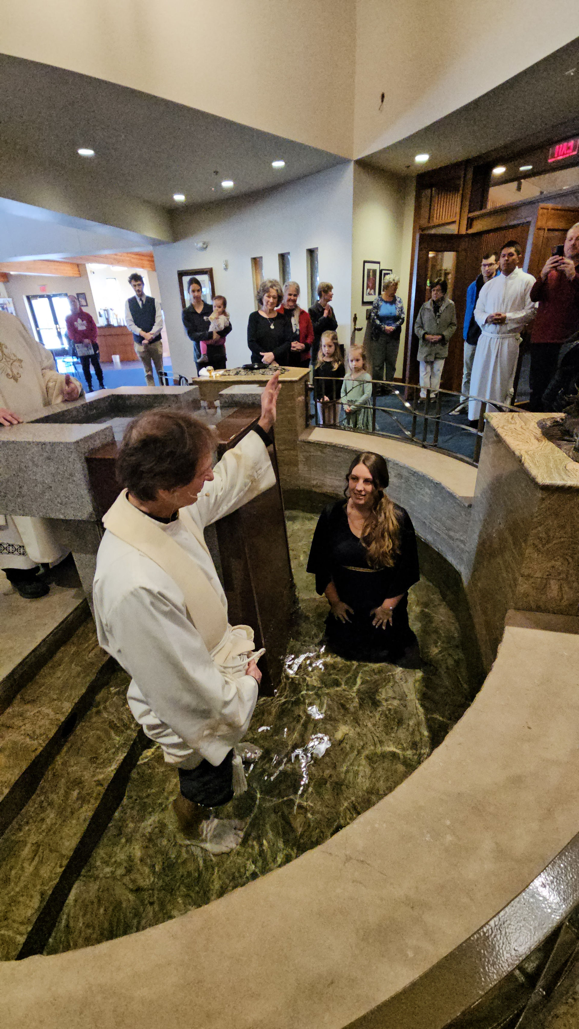 A priest in white robes performs a baptism in a church's water font. A woman kneels in the water, surrounded by onlookers. Warm lighting.