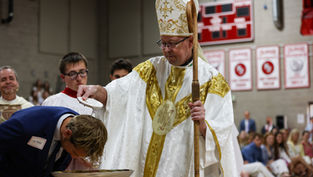 A bishop in ornate robes baptizes a person in a church setting, pouring water over their head. Red banners hang in the background.