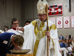 A bishop in ornate robes baptizes a person in a church setting, pouring water over their head. Red banners hang in the background.