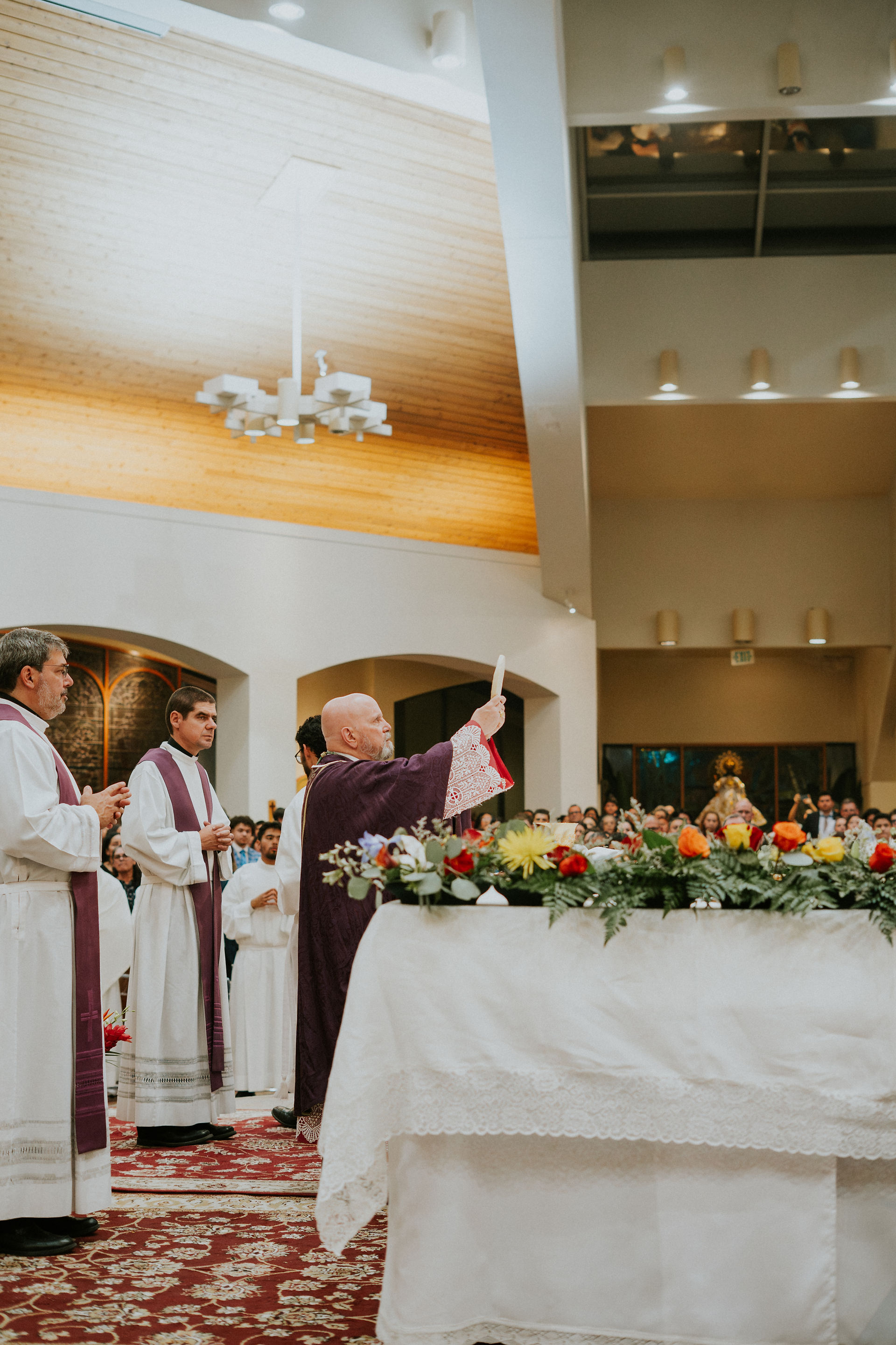 A bishop in purple robes raises an item during a church service, surrounded by clergy in white. A floral display adorns the altar.