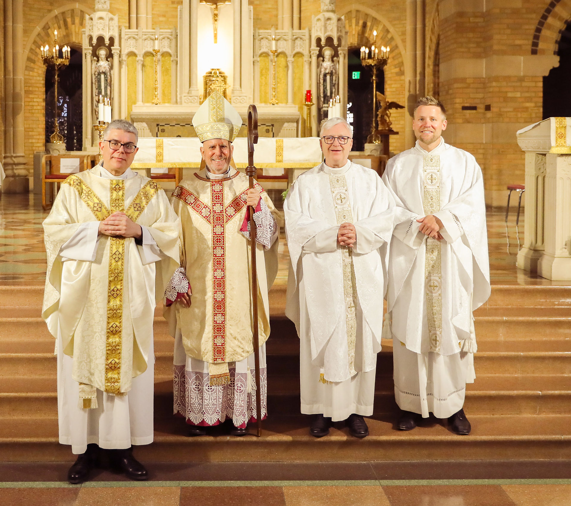 Three priests stand for a photo with an archbishop in an ornate chapel following a religious ceremony.