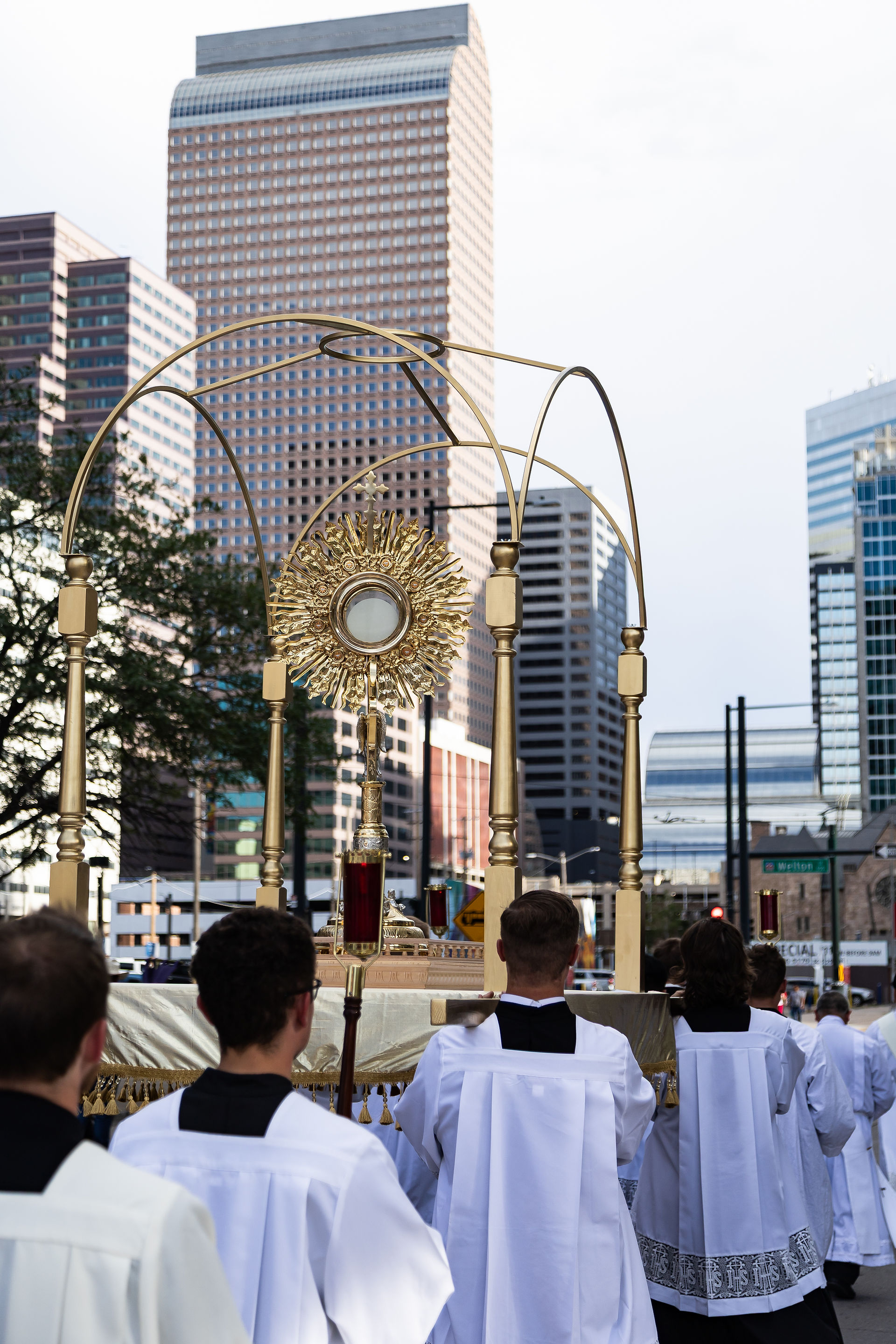 Clergy in white robes process behind a gold monstrance during an outdoor Catholic ceremony, with skyscrapers rising behind.