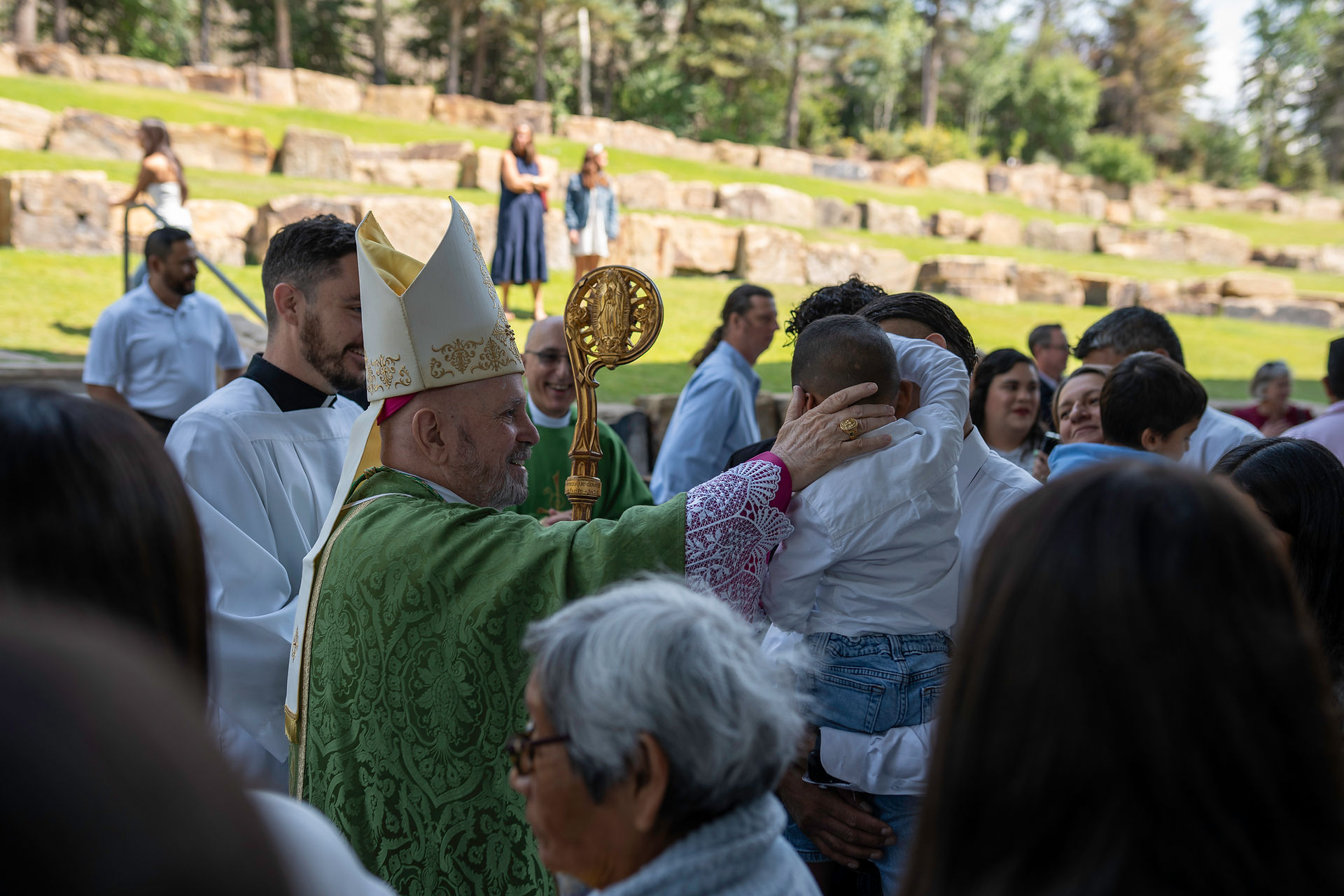 A bishop in green robes blesses a child outdoors. People gather around on a sunny day, with a grassy, rocky background.