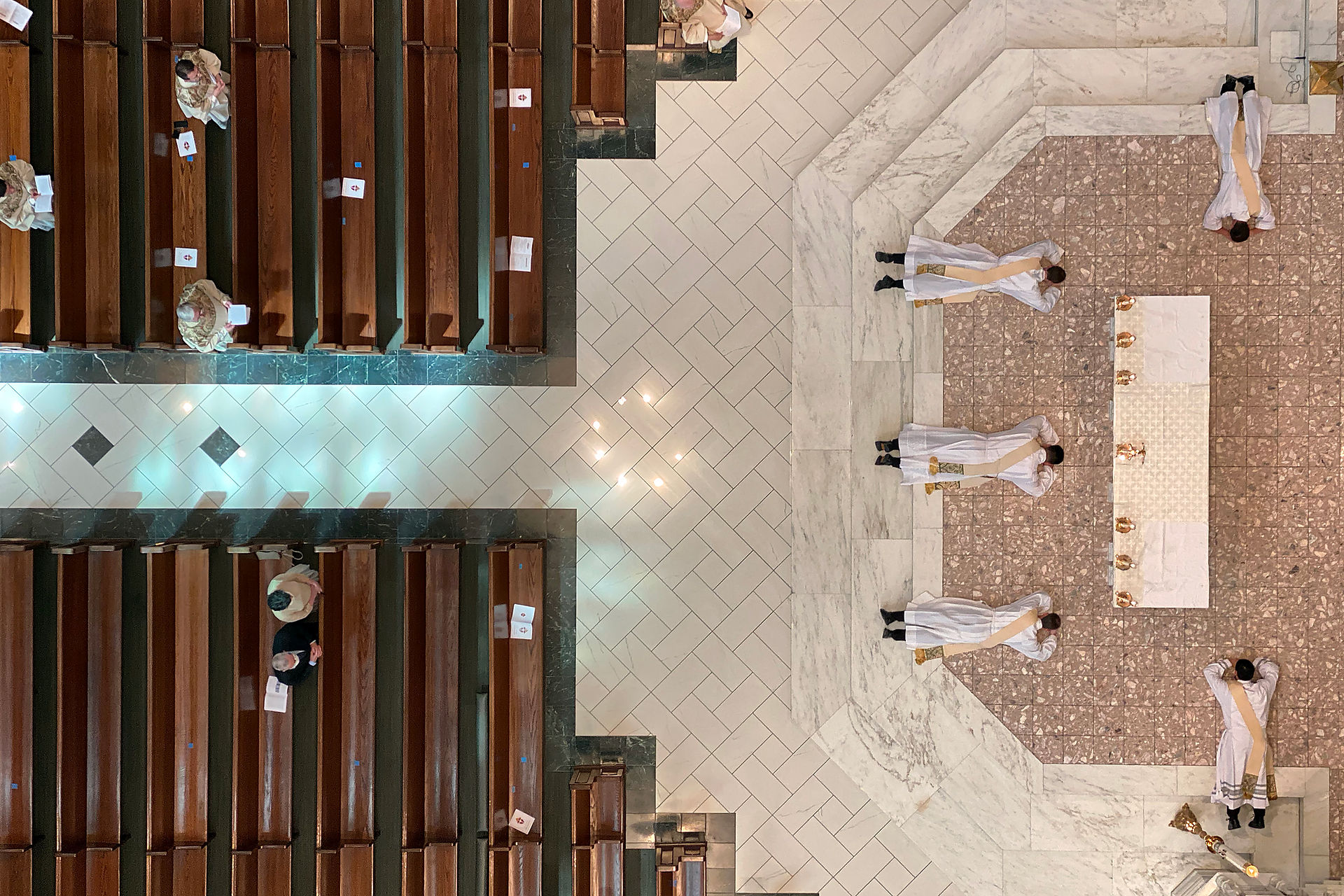Aerial view of a church interior. Priests in white robes lie prostrate before an altar. People sit spaced in wooden pews, creating a solemn mood.